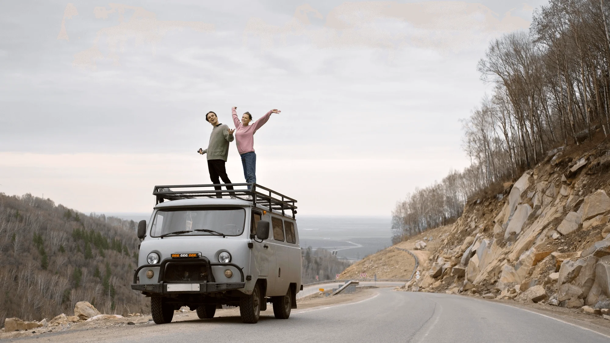 Two people stand on the roof rack of a parked van on a winding road, with trees and rocky hillsides in the background under a cloudy sky.