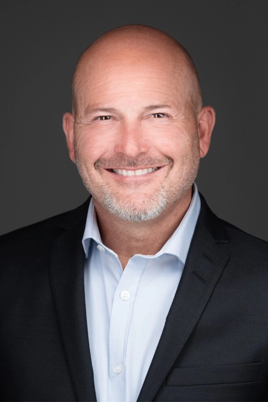 A bald man in a suit jacket and dress shirt smiles at the camera against a plain dark background.
