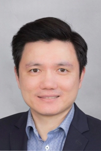 A man with short black hair wearing a dark blazer and light blue button-up shirt, posing against a plain light grey background.