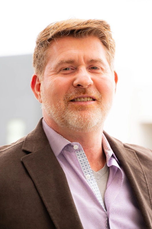 A middle-aged man with short light brown hair and a beard, wearing a brown jacket over a light purple shirt, smiles at the camera against a blurred outdoor background.