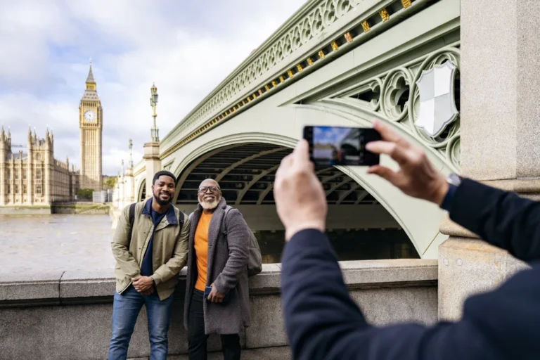 Two men pose for a photo near Westminster Bridge in London, with Big Ben and the Houses of Parliament in the background. Another person takes their picture with a smartphone.